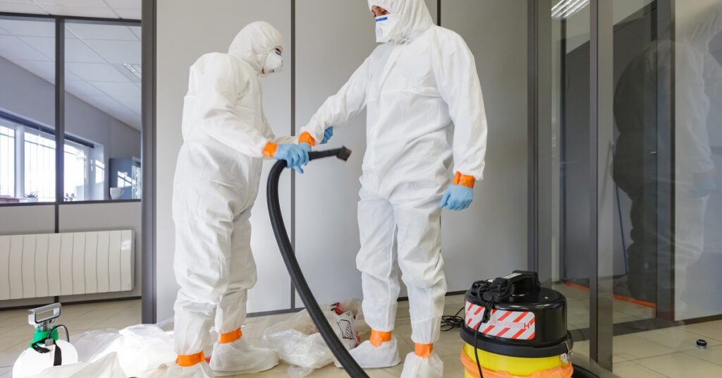 Two people wearing white hazmat suits and holding a vacuum cleaner in a gray room with a tile floor.